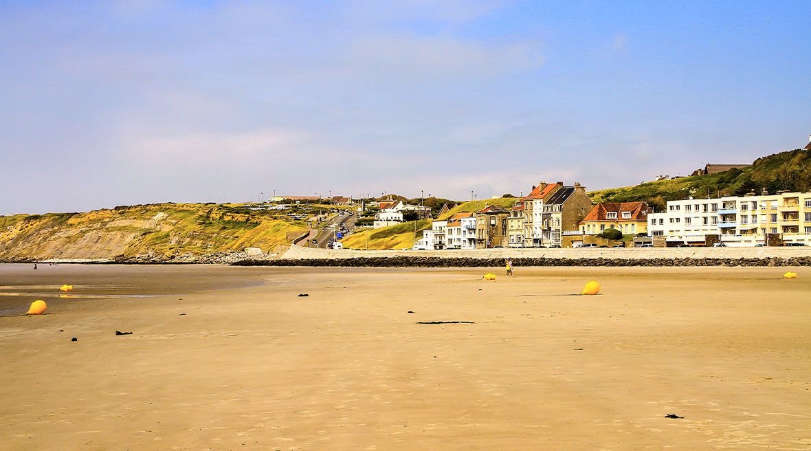Huge stretch of golden sand dotted with yellow buoys with a row of buildings in the distance and cliffs under blue sky
